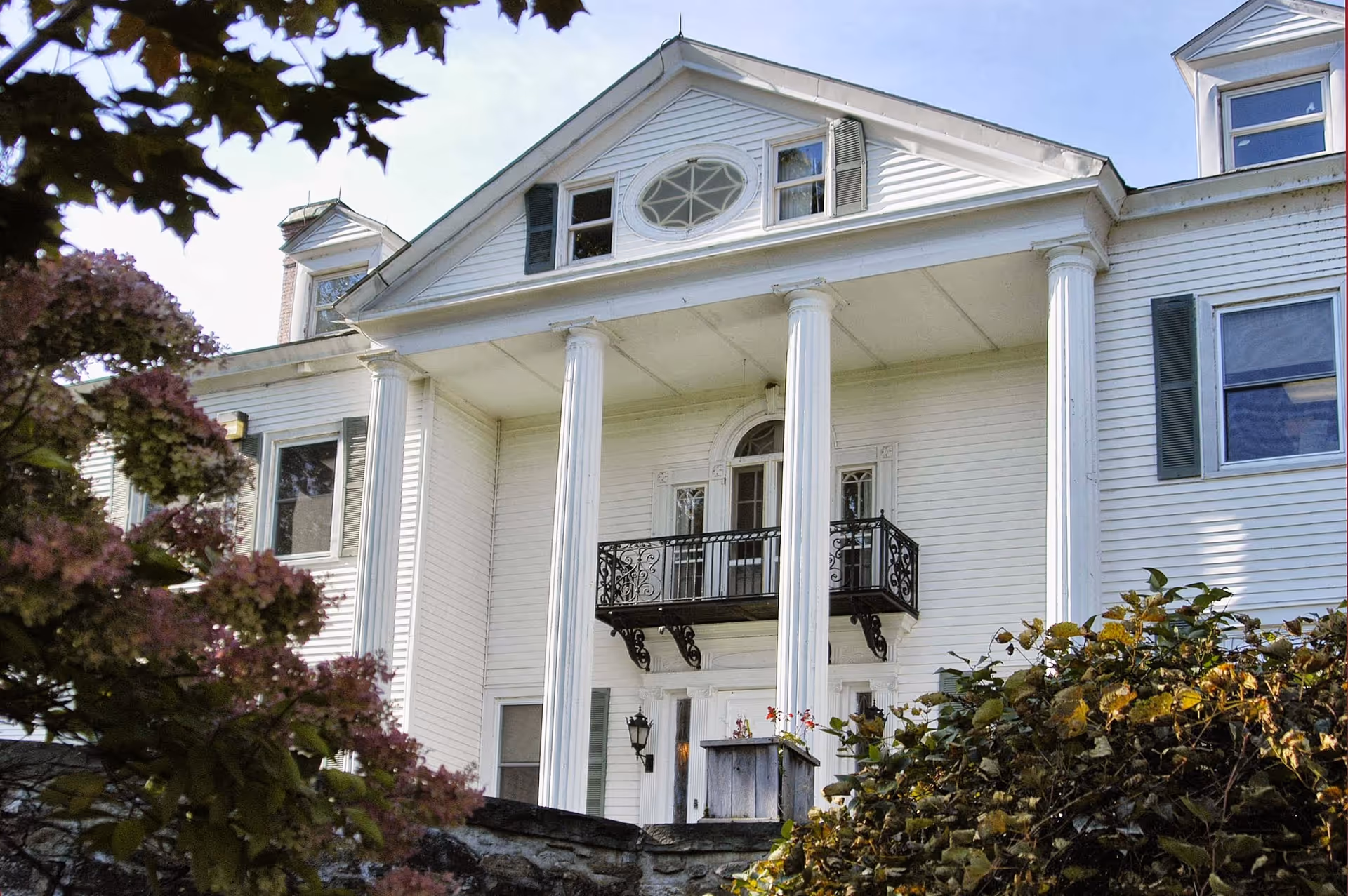 Front exterior view of a large white building with classical architectural features including tall white columns, a small balcony with ornate black railing, and an oval window near the roof. The building is partially obscured by trees and bushes in the foreground.
