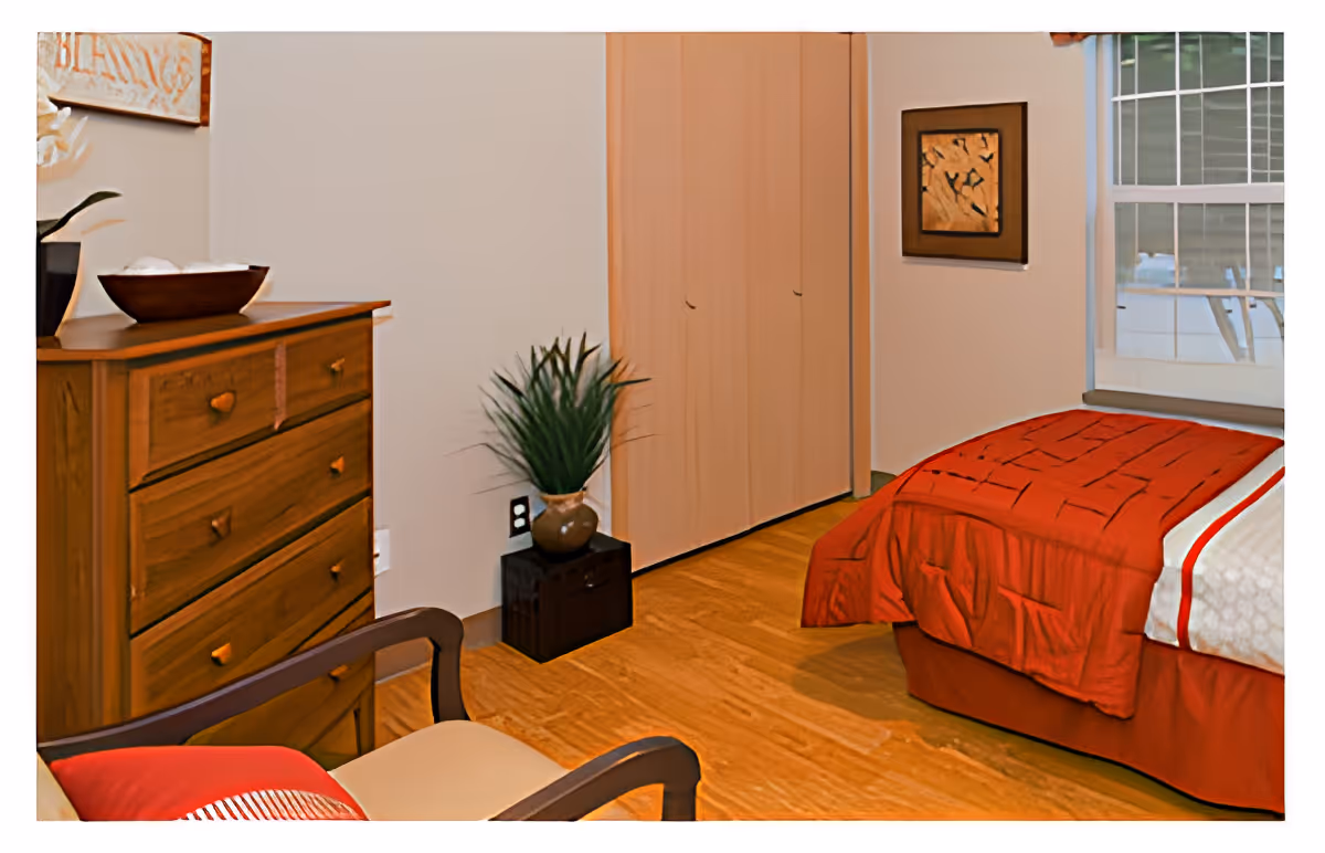 A cozy bedroom featuring a wooden dresser with a decorative bowl and plant on top, a chair with a red cushion, a bed with a red comforter and white sheets, a closet with wooden sliding doors, a framed artwork on the wall, and a window letting in natural light.