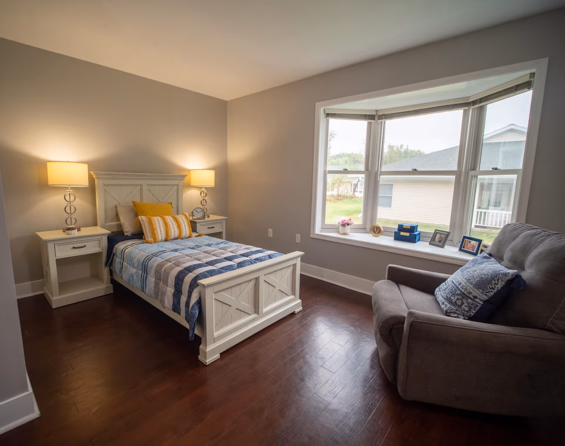 Well-lit bedroom with a white single bed flanked by nightstands and lamps and a cushioned armchair by a bay window.