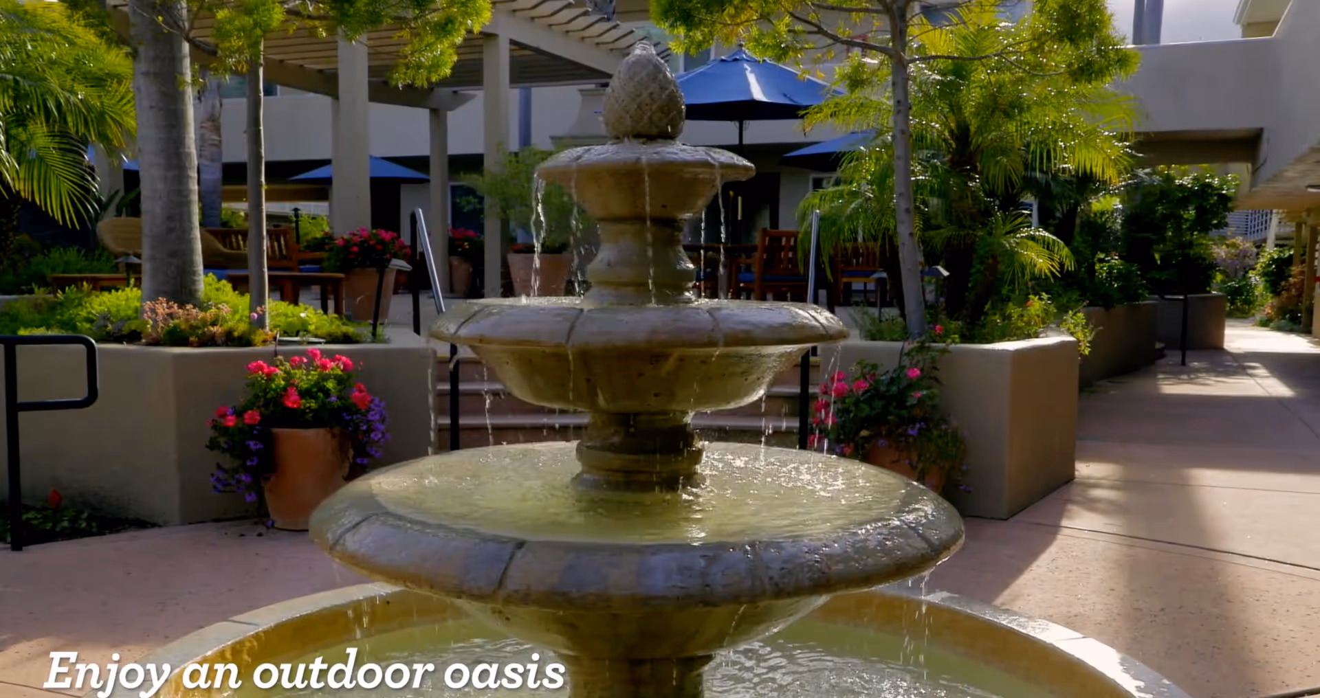 A multi-tiered stone water fountain in the center of a courtyard surrounded by potted plants, trees, and outdoor seating with blue umbrellas at The Village at The Palms.