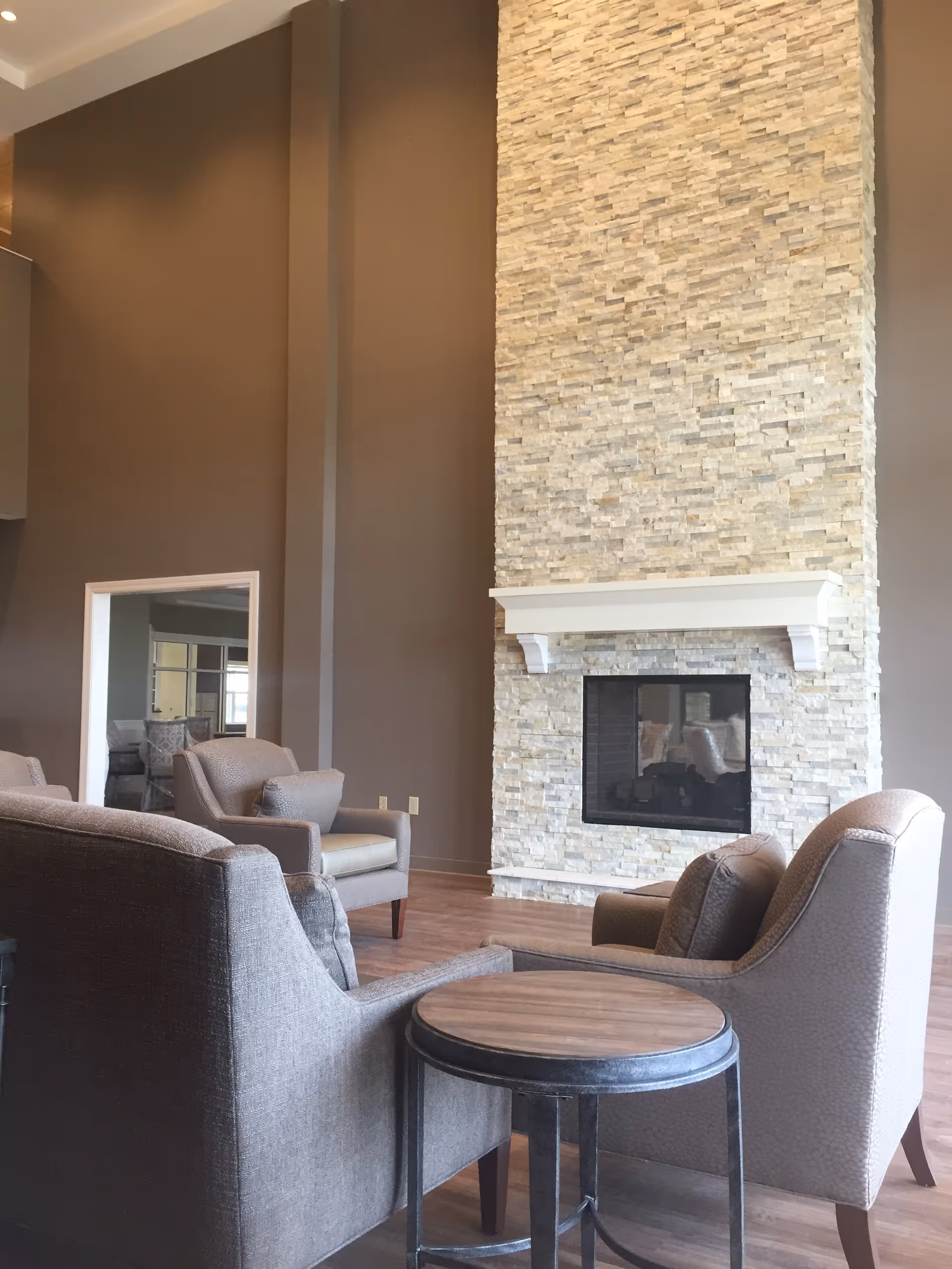 A cozy living room area with a tall stone fireplace as the focal point. The room features neutral-toned armchairs and a sofa arranged around a small round wooden table. The walls are painted in a warm taupe color, and the floor is wood.