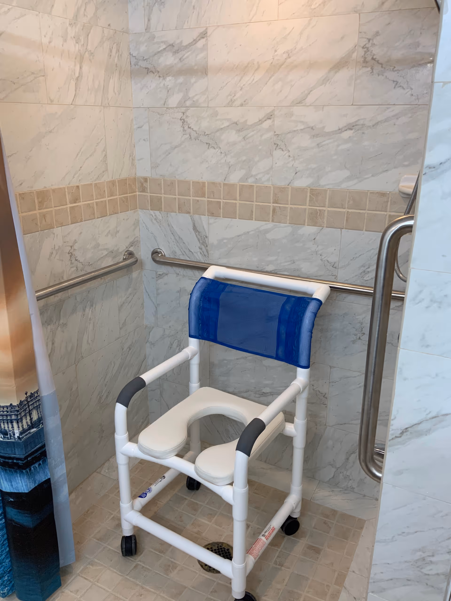 A shower area with marble-patterned tile walls and a beige tiled floor. Inside the shower is a white and blue shower chair with wheels, designed for accessibility. There are stainless steel grab bars mounted on the walls for support and safety.