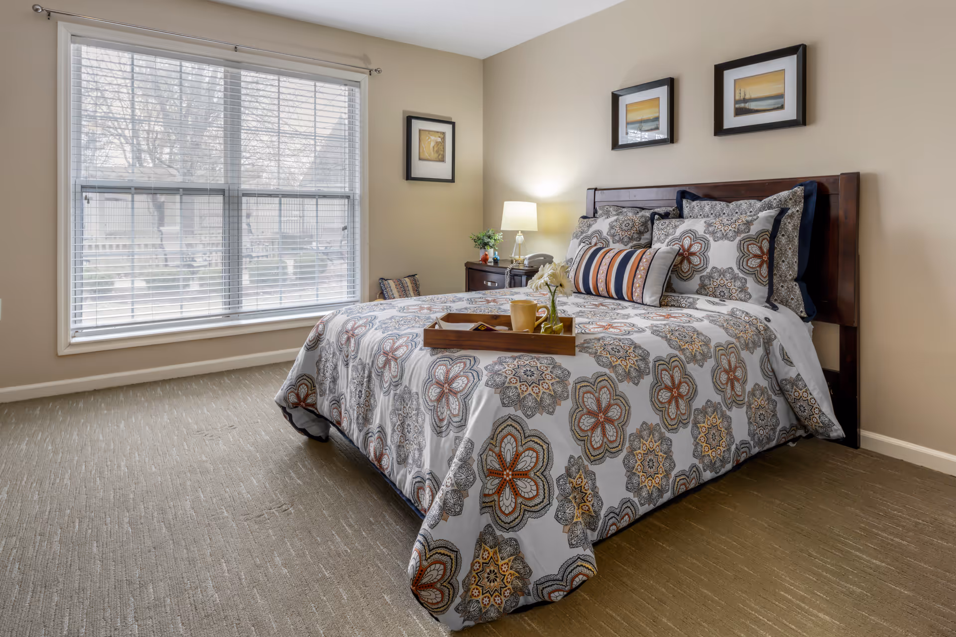A neatly made bed with a floral patterned comforter and multiple pillows in a bedroom. A wooden nightstand with a lamp, a small plant, and a telephone is next to the bed. There are three framed pictures on the beige walls above the bed. A large window with white blinds lets in natural light, and a wooden tray with a cup and flowers is placed on the bed.