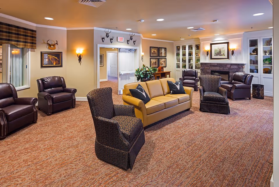 A cozy living room area in a memory care community featuring a tan leather sofa with two decorative pillows, four dark leather armchairs, and two patterned fabric chairs arranged on a carpeted floor. The room has warm lighting with wall sconces, a stone fireplace with a framed deer picture above it, built-in white shelves with decorative items, and several framed animal-themed artworks on the walls. There is a window with plaid valance and an exit door in the background.