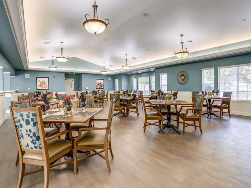 A spacious dining room with multiple wooden tables and chairs arranged neatly. Each table is set with placemats, glassware, and napkins, with small floral centerpieces. The room has light blue walls, large windows allowing natural light, and ceiling lights with decorative fixtures.