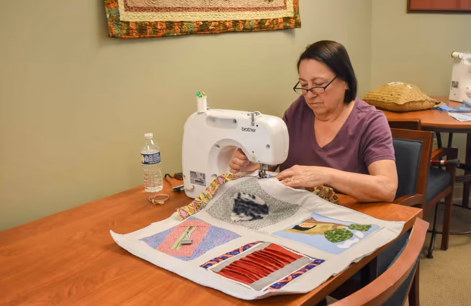 A woman wearing glasses and a purple shirt is sitting at a wooden table using a white Brother sewing machine to work on a colorful quilt. There is a water bottle on the table and another sewing machine and a woven basket on a table in the background. A quilt is hanging on the wall behind her.