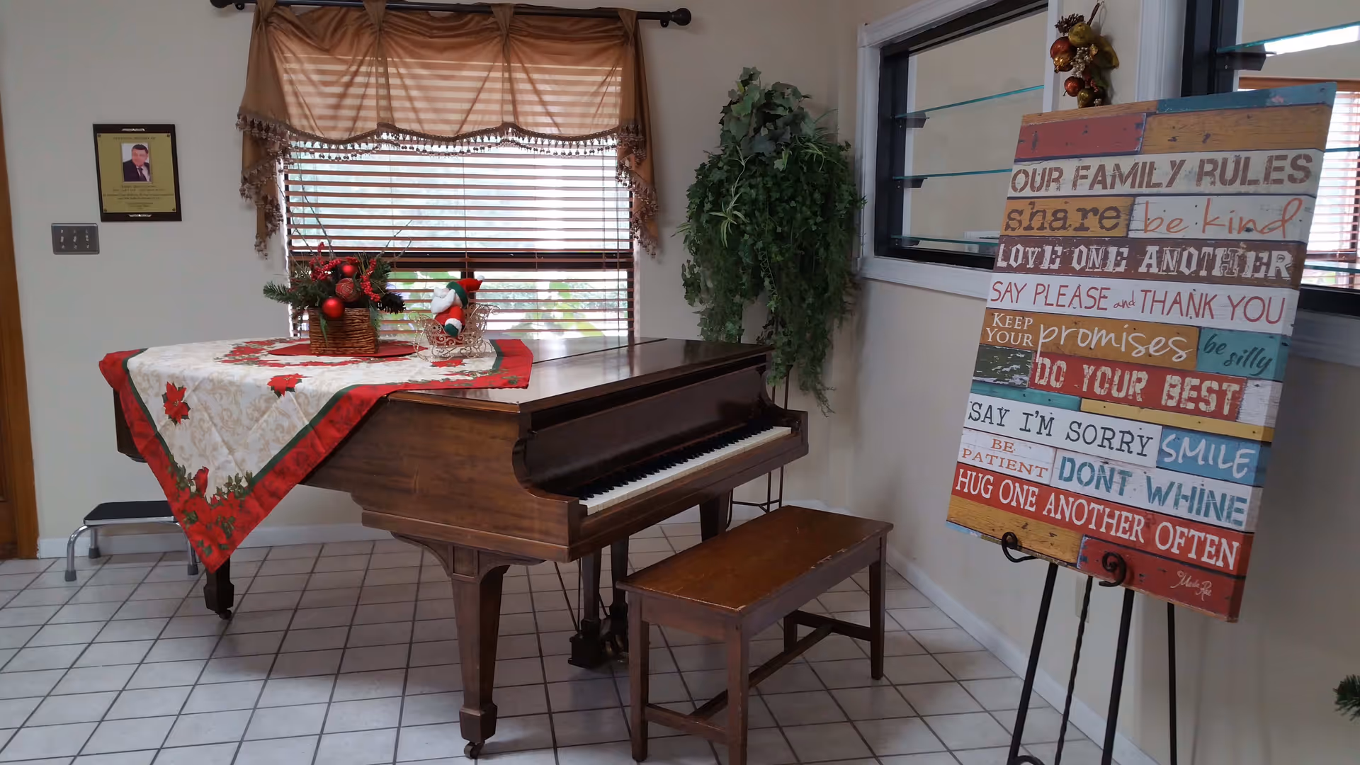 A wooden grand piano with a matching bench is placed on a tiled floor near a window with brown blinds and a valance. The piano is decorated with a festive tablecloth and a small holiday arrangement. To the right of the piano, there is a colorful sign on an easel displaying family rules such as 'share,' 'be kind,' and 'do your best.' A green leafy plant hangs on the wall behind the sign.
