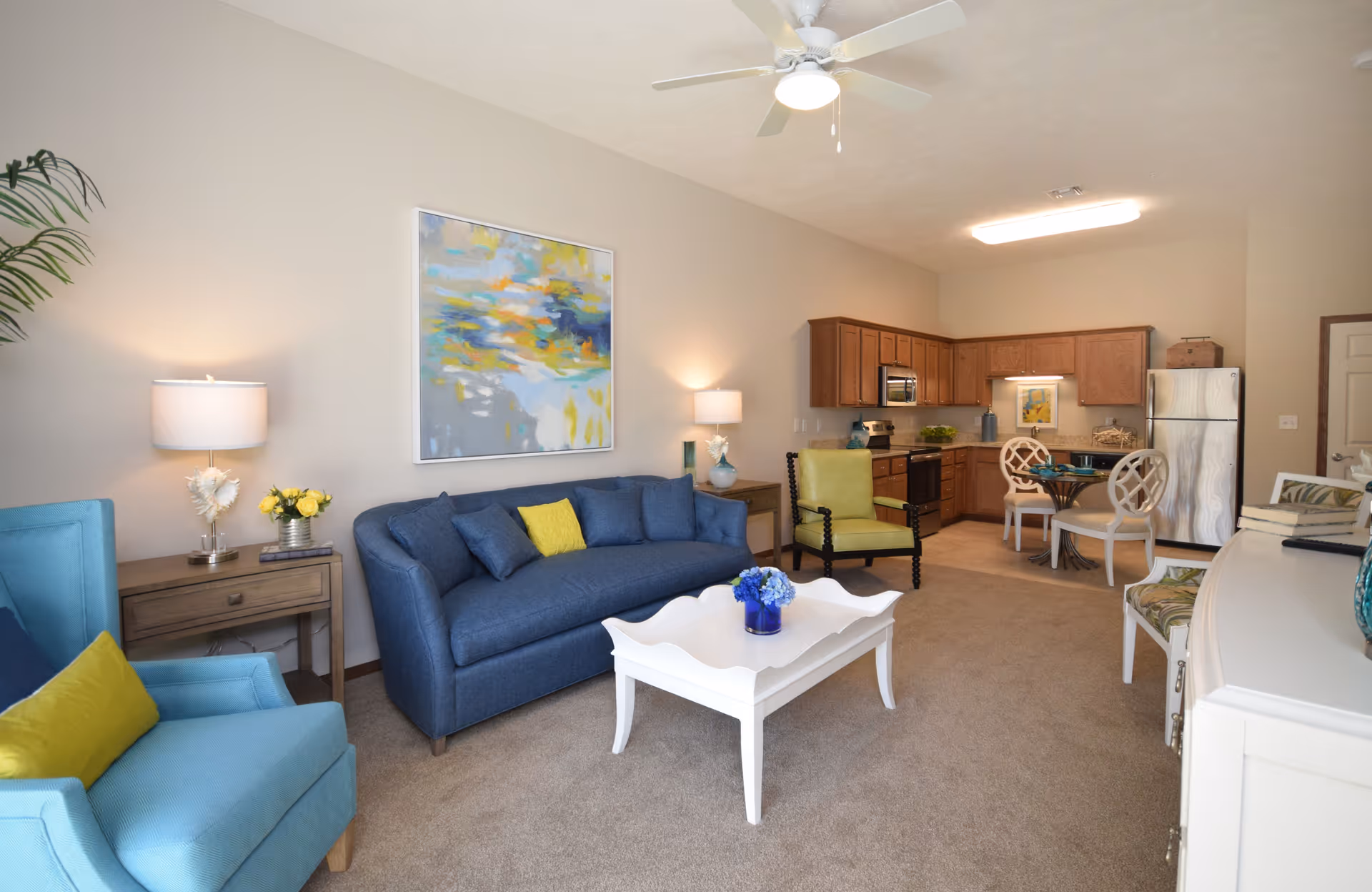 A bright and cozy living area in a retirement community featuring a blue sofa with cushions, a white coffee table with a small flower arrangement, two armchairs with yellow cushions, a side table with a lamp and flowers, a ceiling fan, and an open kitchen with wooden cabinets, stainless steel appliances, and a small dining table with four chairs.