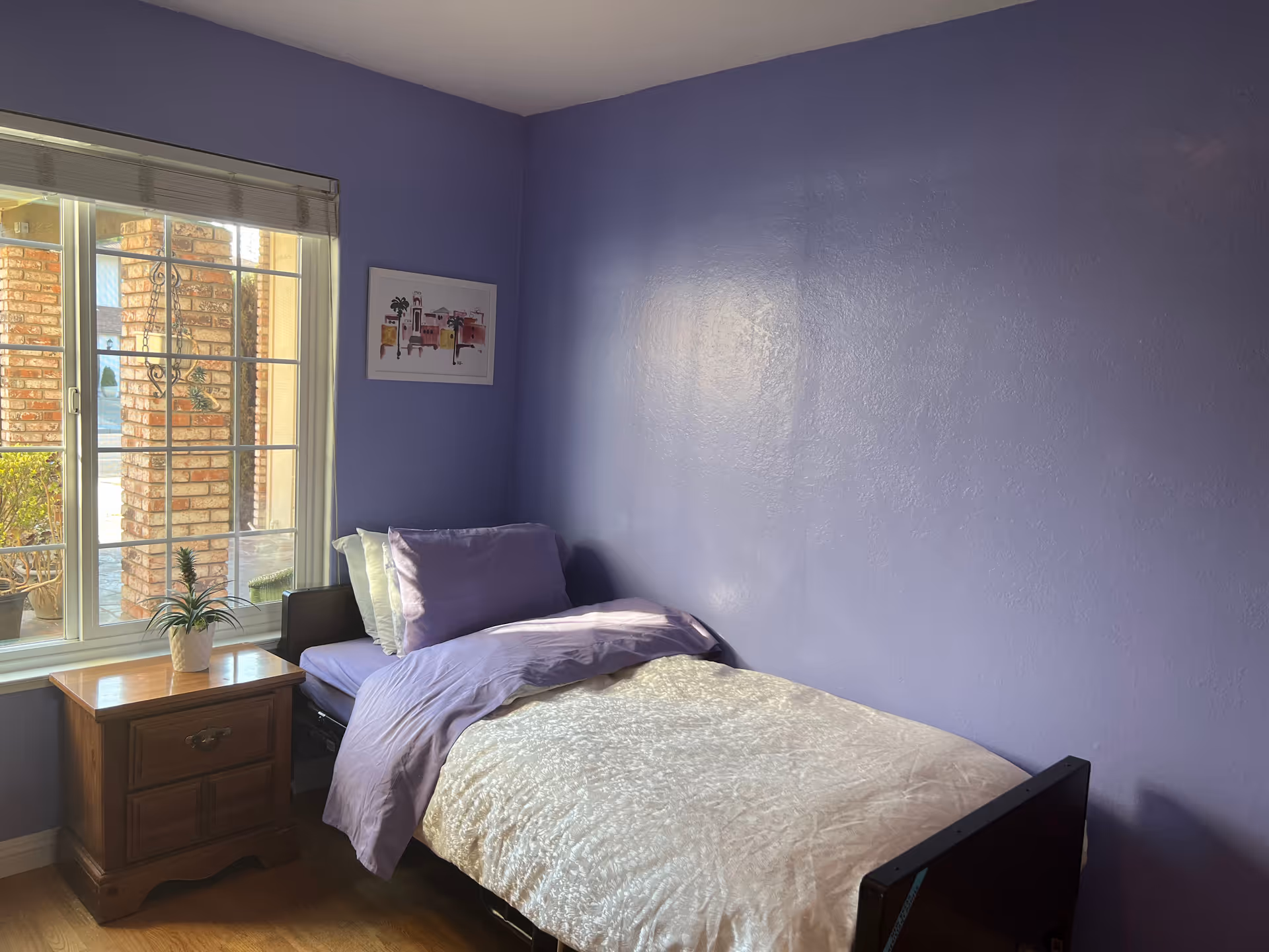 Small bedroom with a single bed against a purple wall, a wooden bedside table by a window, and framed artwork on the wall.