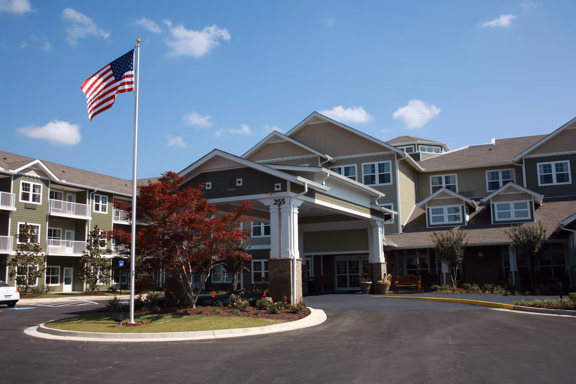 Exterior view of a multi-story senior living facility with a covered entrance, landscaped garden, and an American flag on a flagpole under a blue sky with some clouds.