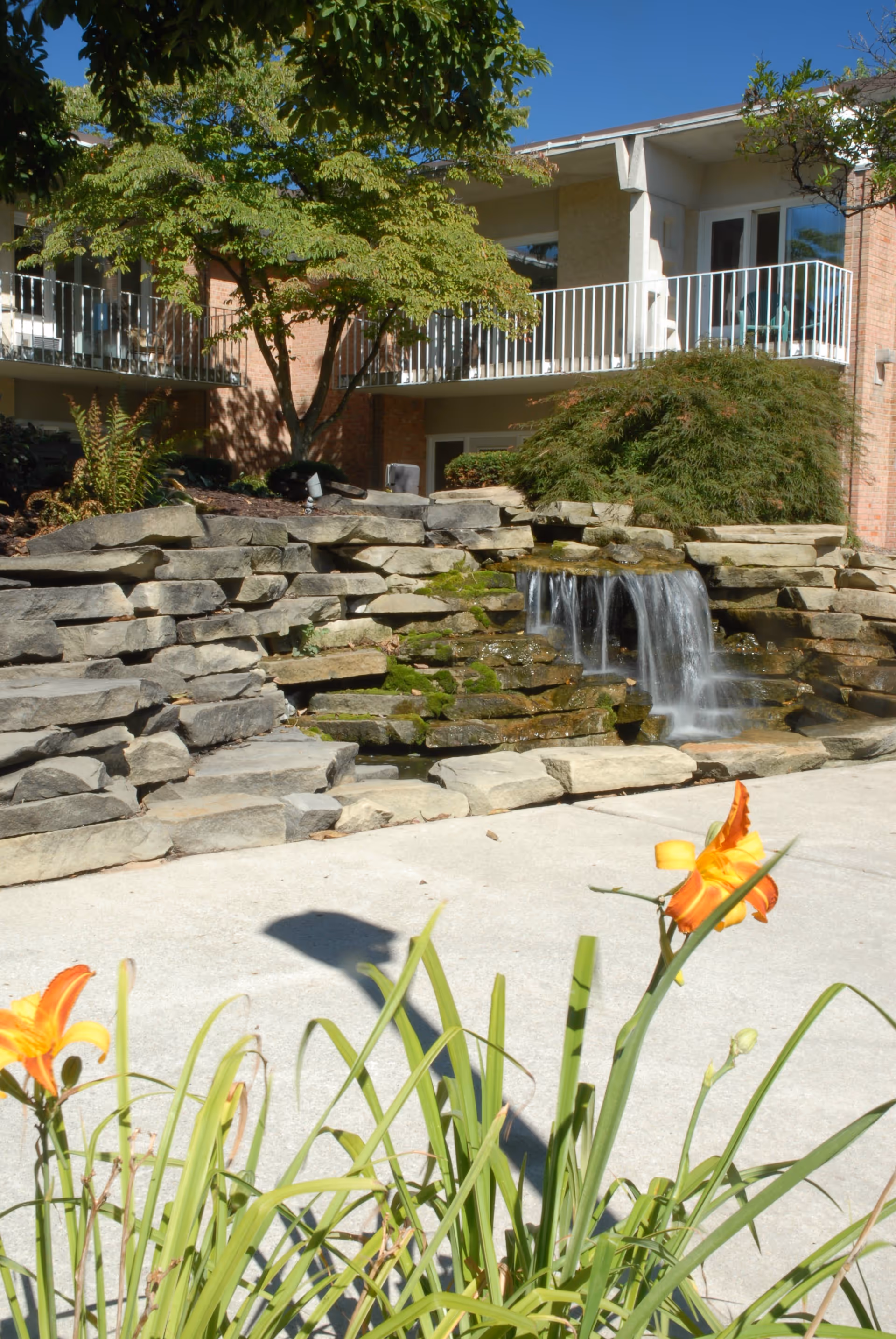 Outdoor courtyard featuring a tiered stone waterfall with orange flowers in the foreground and apartment balconies in the background.