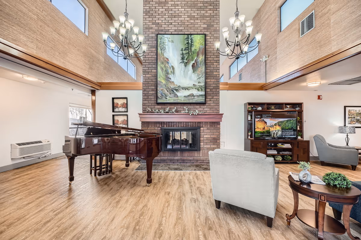 A spacious living room area in a retirement home featuring a grand piano, a brick fireplace with a large painting of a waterfall above it, two chandeliers, comfortable armchairs, a wooden coffee table with decorative items, and a TV on a wooden stand. The room has high ceilings with large windows and wood flooring.