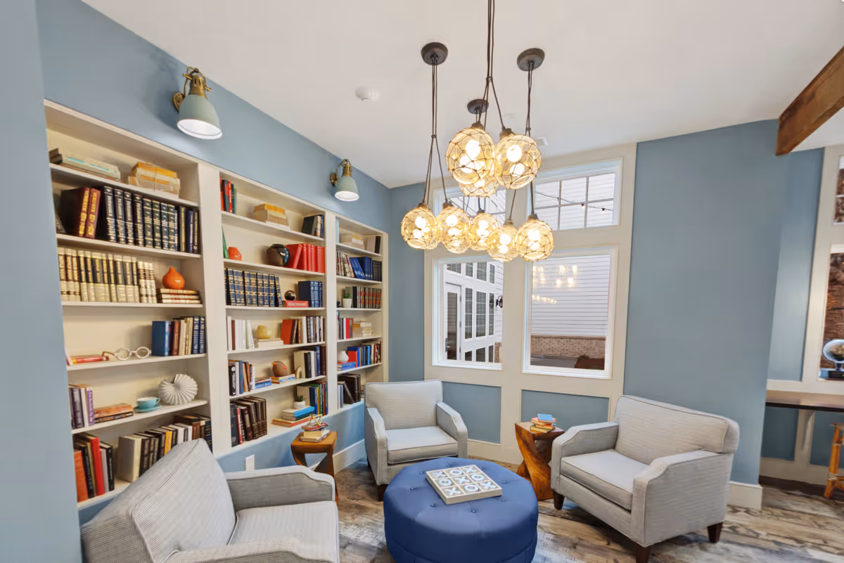 A cozy sitting area with three gray armchairs arranged around a blue ottoman with a tic-tac-toe game on top. The walls are painted blue and feature built-in white bookshelves filled with books and decorative items. Above the seating area hangs a modern chandelier with multiple globe-shaped lights. Large windows let in natural light, and the floor has a wood-like pattern.