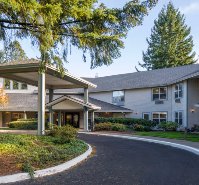Exterior view of a two-story senior living facility named Markam House with a covered entrance, surrounded by trees and landscaped greenery under a clear sky.