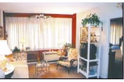 A cozy living room with a large window covered by sheer curtains, a sofa, an armchair with floral upholstery, a small glass coffee table, a white shelving unit with a TV and decorative plants, and a floor lamp.