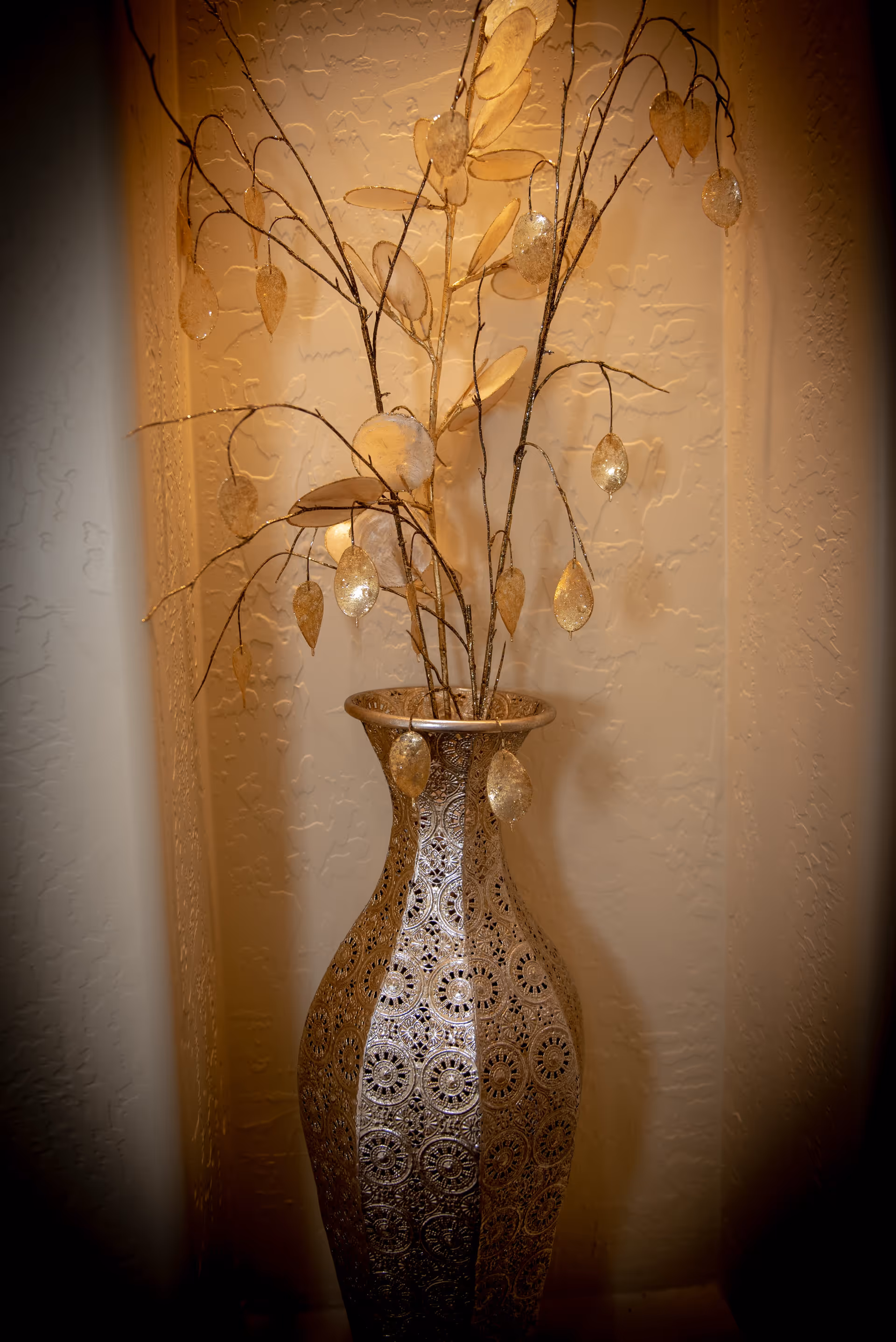 Ornate tall metal vase holding decorative branch stems with gold leaf-like ornaments in a textured wall niche.