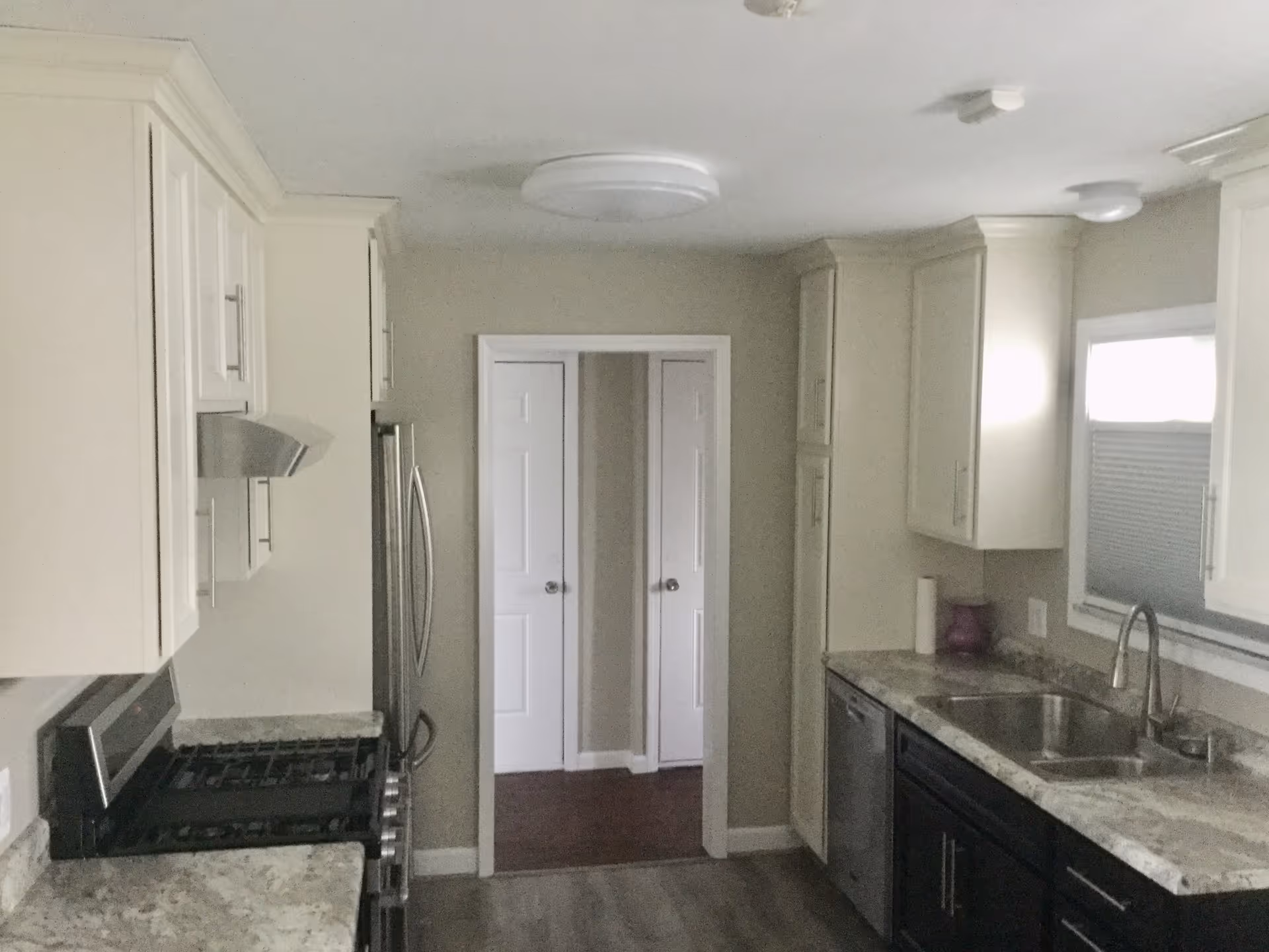 Galley kitchen with stainless steel appliances, white upper cabinets, dark lower cabinets, a double sink under a window, and a doorway at the far end.