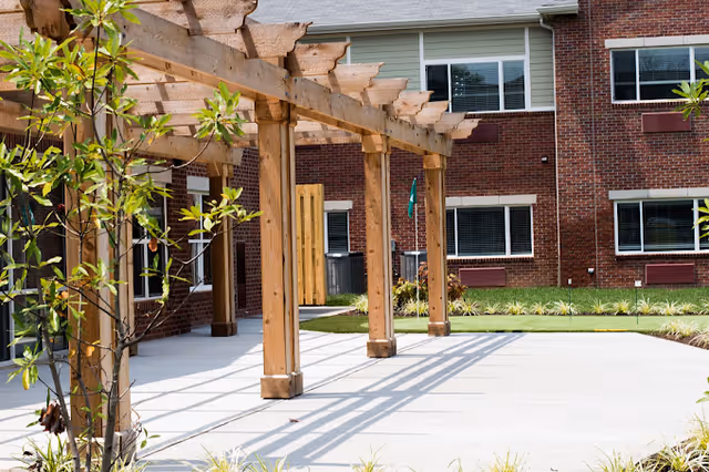 Outdoor patio area with a wooden pergola casting shadows on a concrete surface, surrounded by greenery and a brick building with multiple windows in the background.