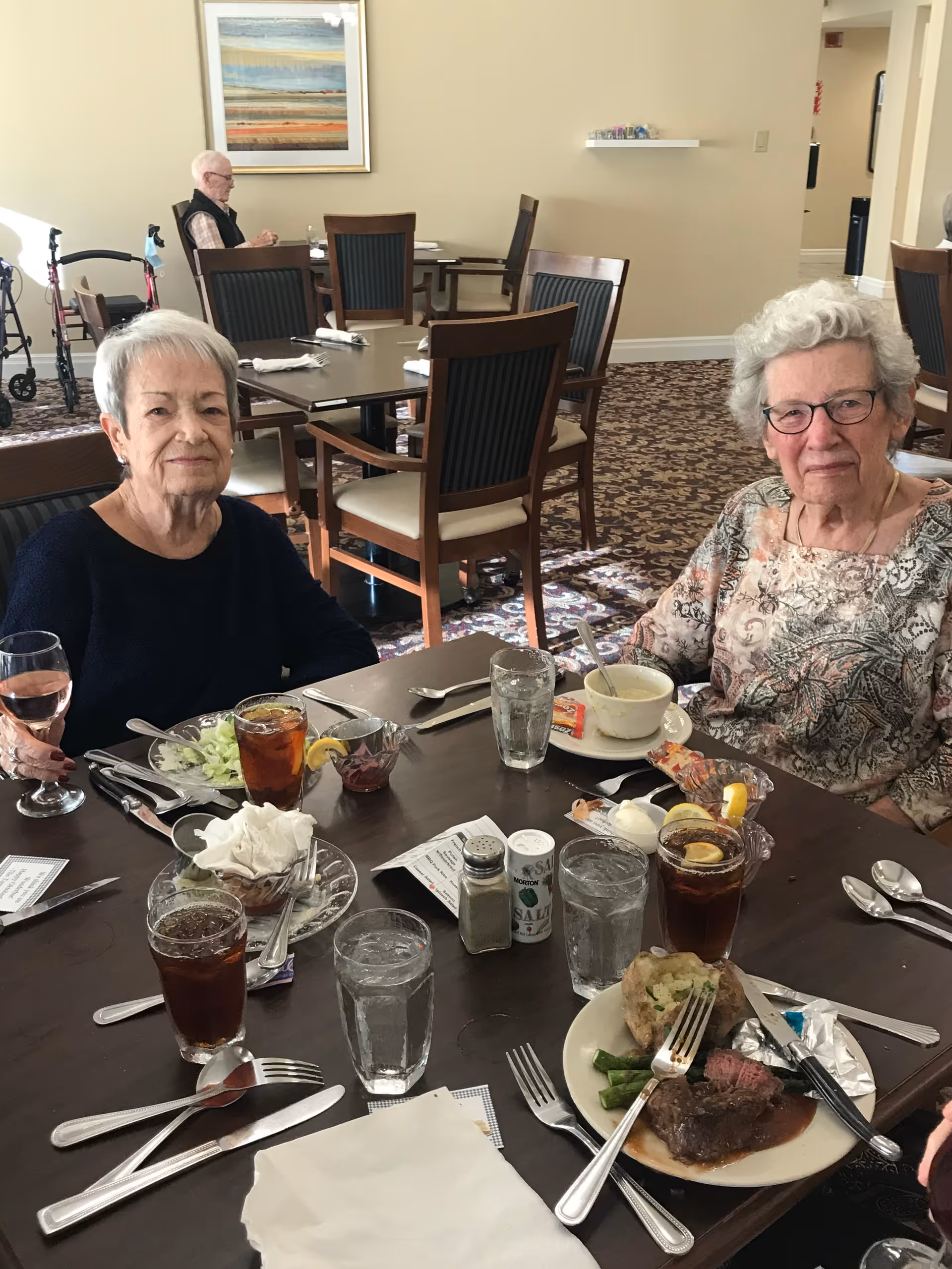 Two elderly women sitting at a dining table in a senior living facility, enjoying a meal with plates of food, glasses of iced tea, and water. The room has several empty tables and chairs, a patterned carpet, and a framed painting on the wall. Another elderly man is seated at a table in the background.