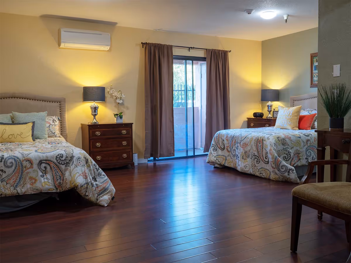 A spacious bedroom with two beds featuring patterned bedspreads and decorative pillows. Each bed has a wooden nightstand with a lamp and small decorative items. There is a sliding glass door with brown curtains leading to an outdoor area. The room has wooden flooring and neutral-colored walls.