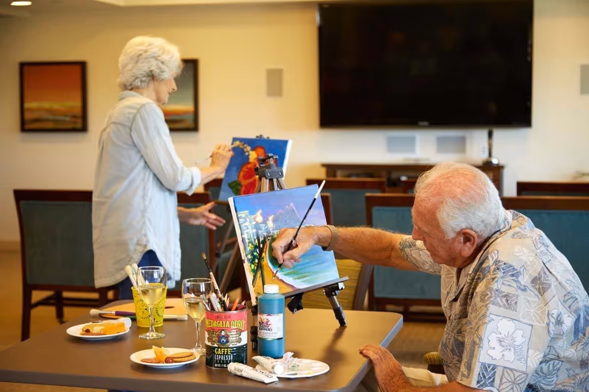 An elderly man and woman painting on canvases set on easels in a well-lit room with chairs and a large TV mounted on the wall. The man is seated at a table with painting supplies, snacks, and drinks, focused on his artwork depicting a beach scene. The woman stands in the background working on her painting.