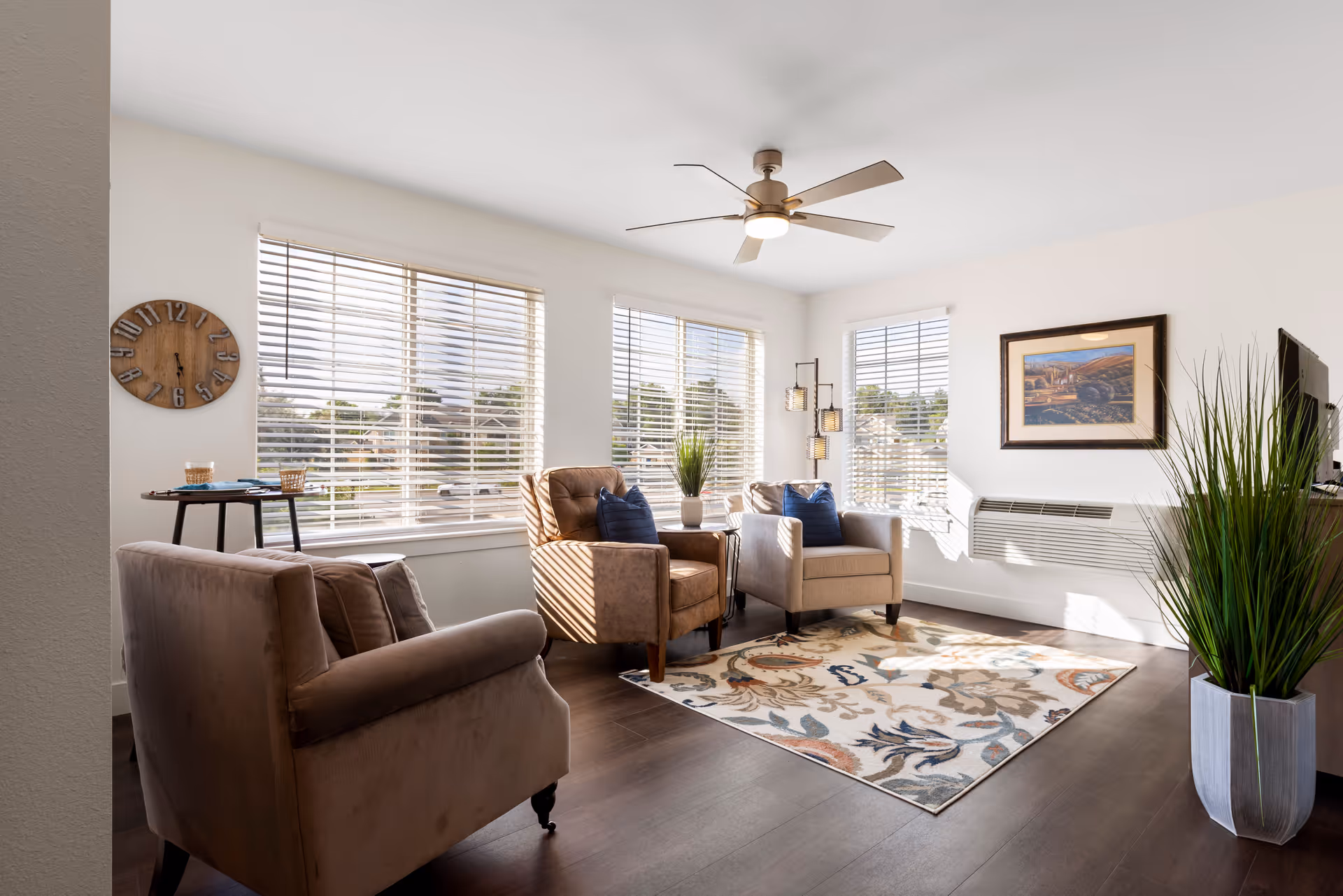 A bright living room with three armchairs arranged around a patterned area rug. Large windows with blinds allow natural light to fill the space. A ceiling fan with a light fixture is mounted on the white ceiling. A wall clock, a framed picture, a floor lamp with three lights, and a tall potted plant add decorative touches to the room.