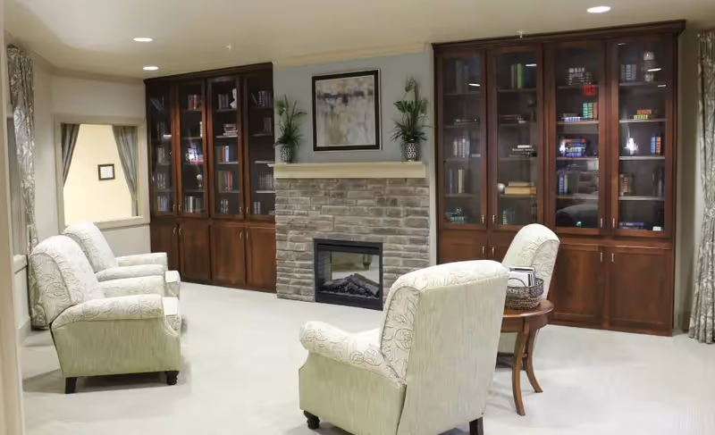 A cozy living room area with four upholstered armchairs arranged around a small wooden side table. The room features a stone fireplace with a mantel decorated with two potted plants and a framed abstract painting above it. On either side of the fireplace are tall wooden cabinets with glass doors, filled with books and decorative items. The room has light-colored carpet and walls, with curtains framing a window on the left side.
