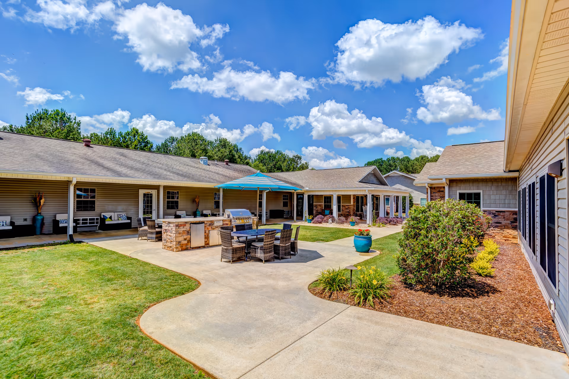 Outdoor courtyard area of a senior living facility with a paved walkway, green lawn, patio furniture including a table with chairs under a blue umbrella, and a built-in grill. The buildings surrounding the courtyard have beige siding and multiple windows. The sky is bright blue with scattered white clouds.