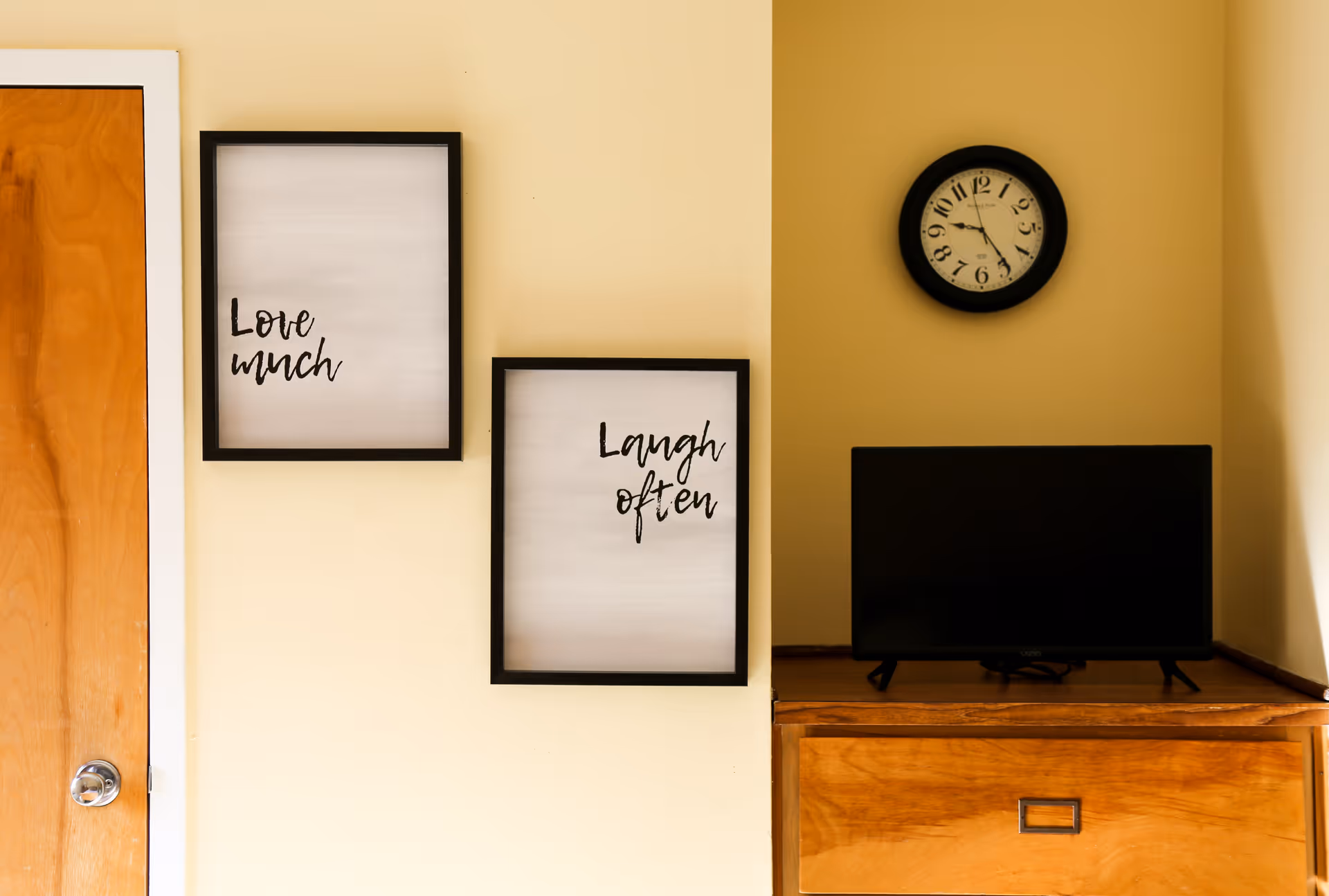 Interior wall with two framed quote prints, a round clock, a TV on a wooden dresser, and a wooden door.
