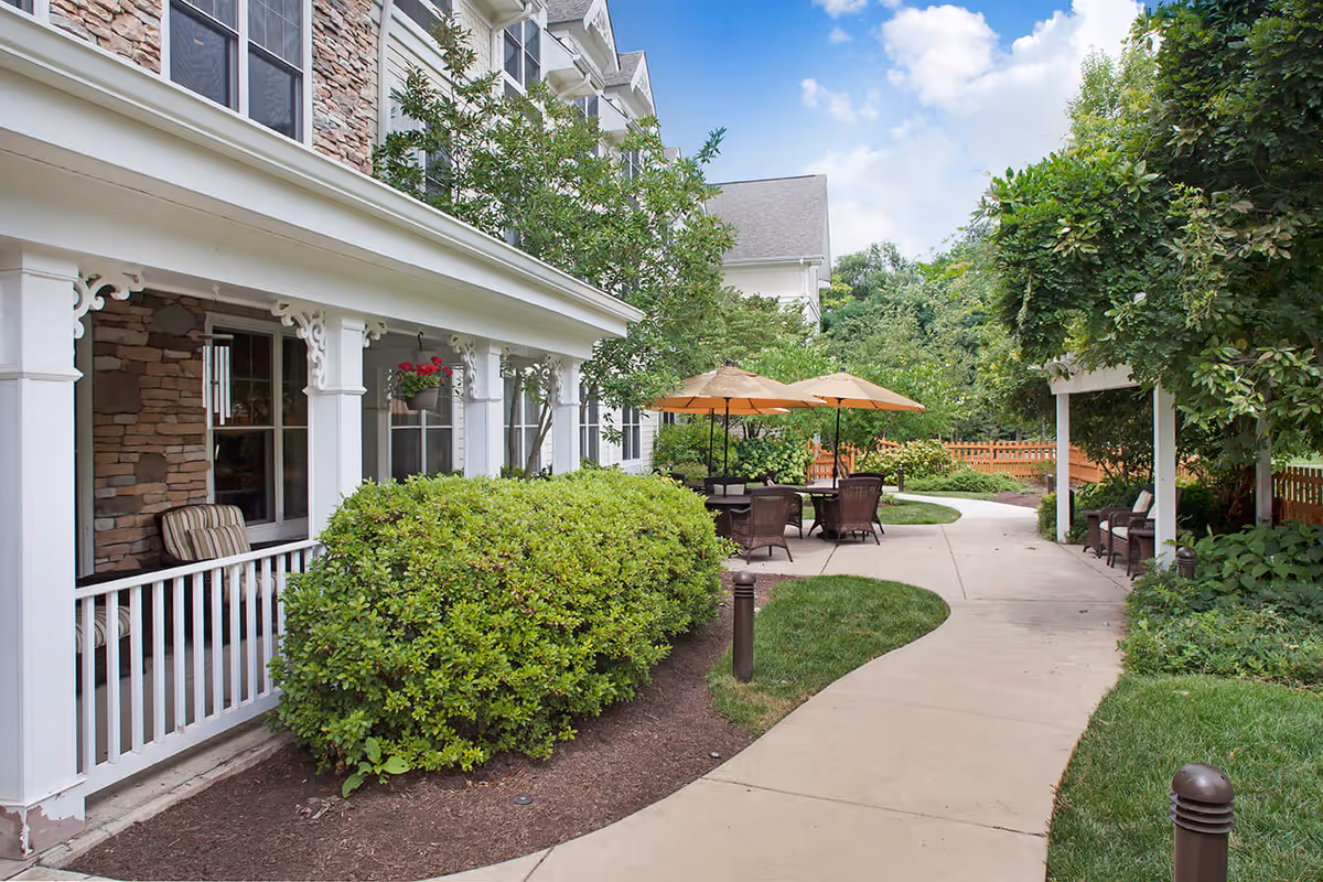 Outdoor patio area at a senior living facility with a winding concrete pathway, green bushes, trees, and seating areas with tables and umbrellas. The building exterior features stone and white trim with windows and a porch with chairs.