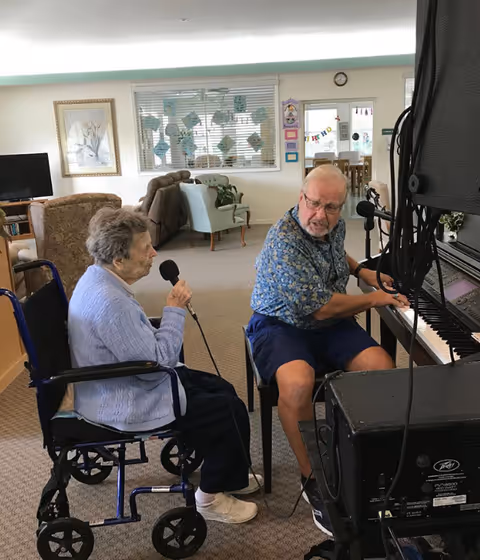 An elderly woman in a wheelchair holding a microphone and an elderly man playing a piano in a common area of an assisted living facility. The room has chairs, a television, and decorations on the walls and windows.