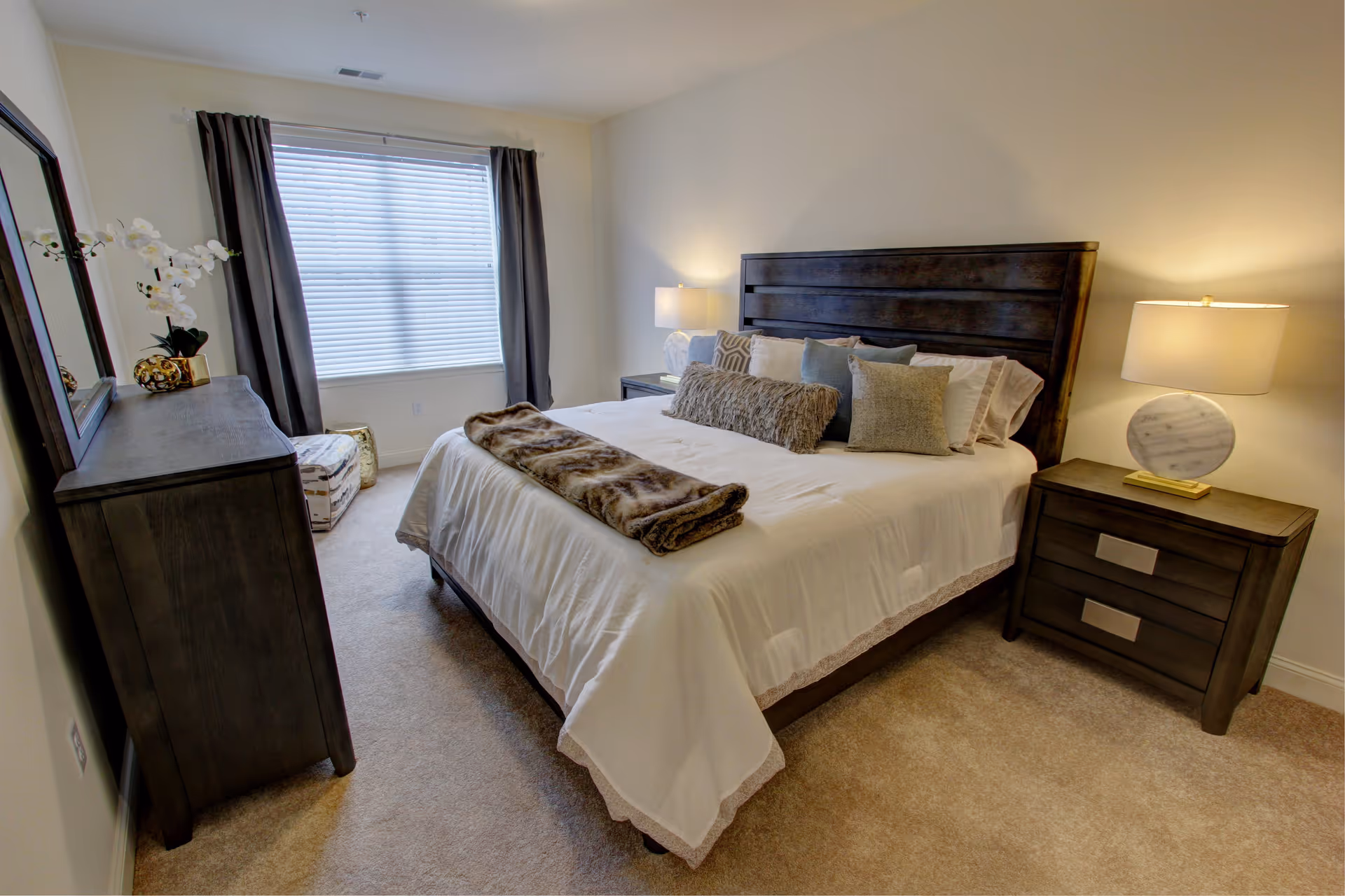 A neatly arranged bedroom featuring a large bed with white bedding and multiple decorative pillows. A brown fur throw is folded at the foot of the bed. On the right side of the bed is a dark wooden nightstand with a modern lamp. On the left side of the room is a matching dark wooden dresser with a large mirror above it and a white orchid plant on top. The room has beige carpet flooring, light-colored walls, and a window with closed blinds and dark curtains.