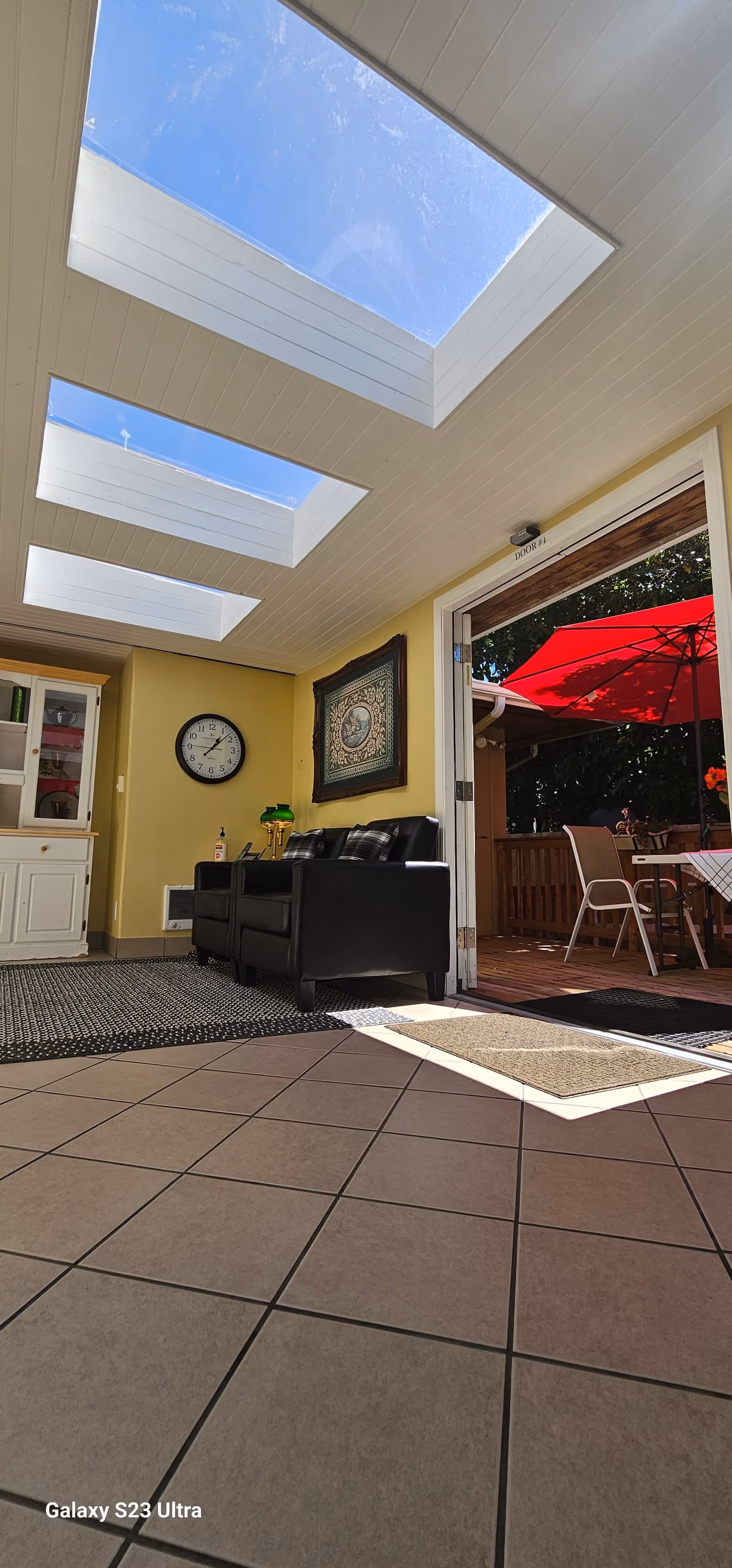 Interior view of a room with large skylights on the ceiling allowing natural light to brighten the space. The room has tiled flooring and a black leather couch against a yellow wall. A clock and framed artwork hang on the wall. To the right, a door opens to an outdoor patio area with a red umbrella and outdoor seating.