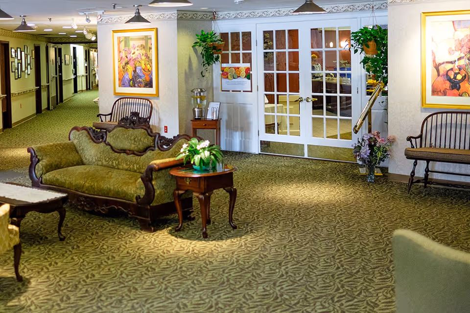 A cozy interior common area with vintage-style green upholstered sofa and wooden chairs. There are framed paintings on the walls, potted plants, and a small wooden side table with a plant on it. Double glass doors lead to another room with tables and chairs visible inside. The carpet has a leafy pattern, and the hallway extends to the left with more framed pictures on the walls.