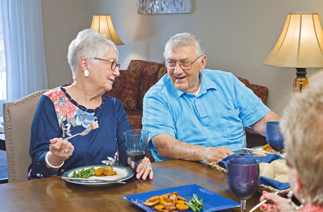 Two elderly people sitting at a dining table, enjoying a meal and engaging in conversation. The woman on the left is holding a fork with food, and the man on the right is smiling. The table has plates with food, glasses, and a basket of bread rolls. The background shows a lamp and a couch.
