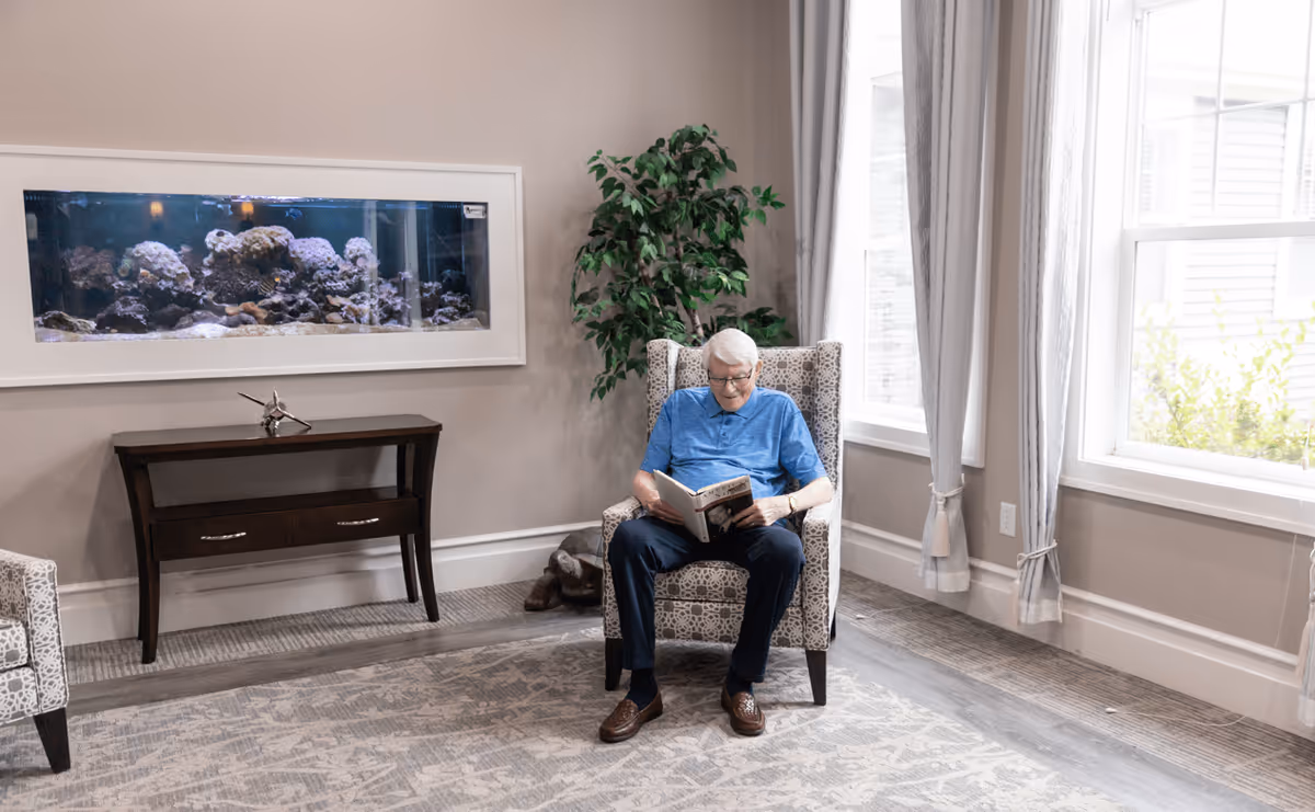 An elderly man with white hair wearing a blue shirt and dark pants is sitting in a patterned armchair reading a book in a well-lit room. Behind him is a large window with light gray curtains, a green potted plant, and a wall-mounted aquarium filled with coral and fish. A small dark wooden table with a decorative object is placed under the aquarium.