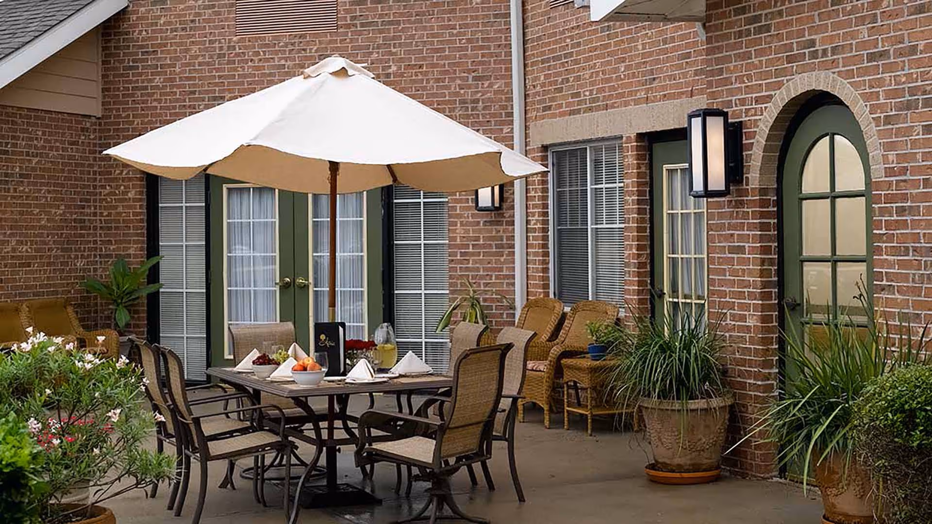 Outdoor patio area with a round table set for dining under a large white umbrella. The table has six chairs around it and is set with napkins, a bowl of fruit, and a pitcher of lemonade. The patio is adjacent to a brick building with green-framed windows and doors. There are potted plants and wicker chairs along the wall.
