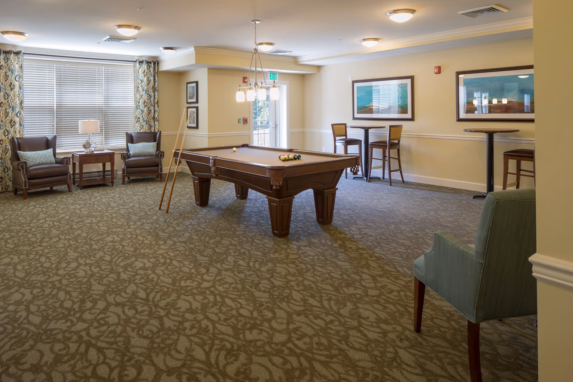 A spacious game room with a pool table in the center, two pool cues resting against the table, two leather armchairs with a small table and lamp between them near a window with patterned curtains, two high tables with chairs along the wall, framed artwork on the walls, and a single upholstered chair in the foreground.