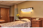 A single bed in a nursing facility room with a floral bedspread, a wooden nightstand with a telephone, a vase of yellow flowers on a small table, a wall-mounted light fixture above the bed, and a closed wooden door in the background.