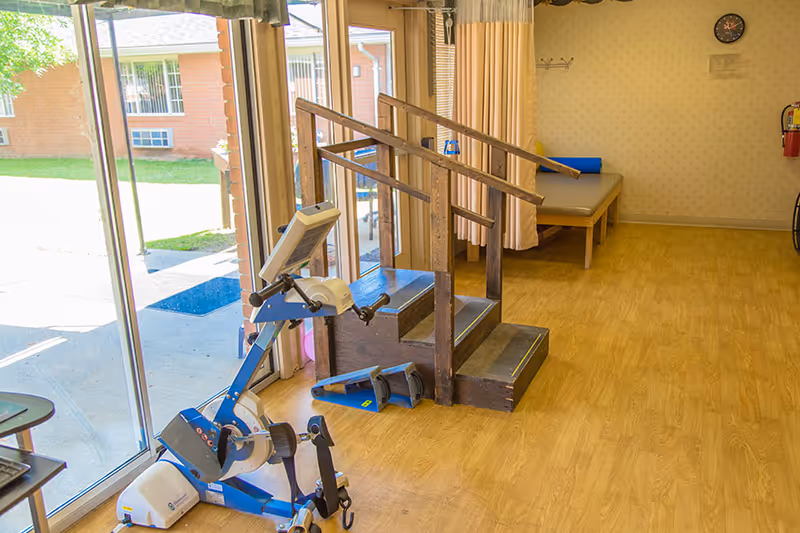 A rehabilitation room with wooden flooring featuring a small set of wooden stairs with handrails for physical therapy. There is exercise equipment including a stationary pedal exerciser near large glass sliding doors that open to an outdoor area with grass and a brick building. A padded bench and a fire extinguisher are visible against the wall inside the room.