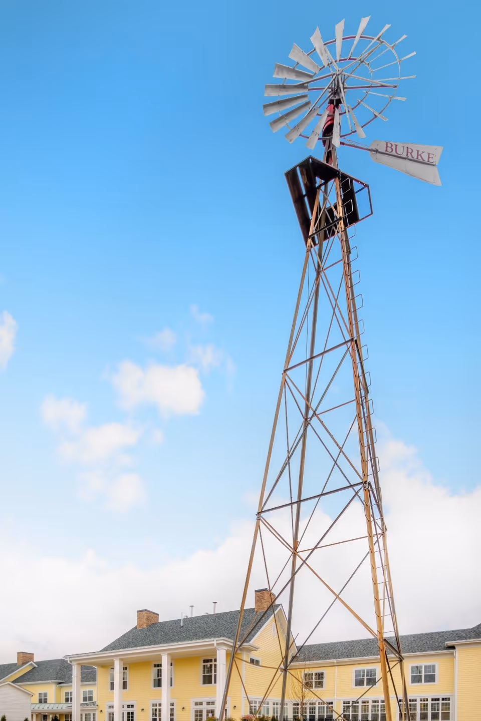 Tall metal windmill structure with the word 'BURKE' on the tail, standing in front of a large yellow building with white trim and multiple windows under a blue sky with some clouds.