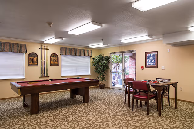 A recreational room with a pool table and a small table with four chairs. The room has patterned carpet, beige walls, two windows with valances, framed artwork, a potted plant near a glass door leading outside, and ceiling fluorescent lights.