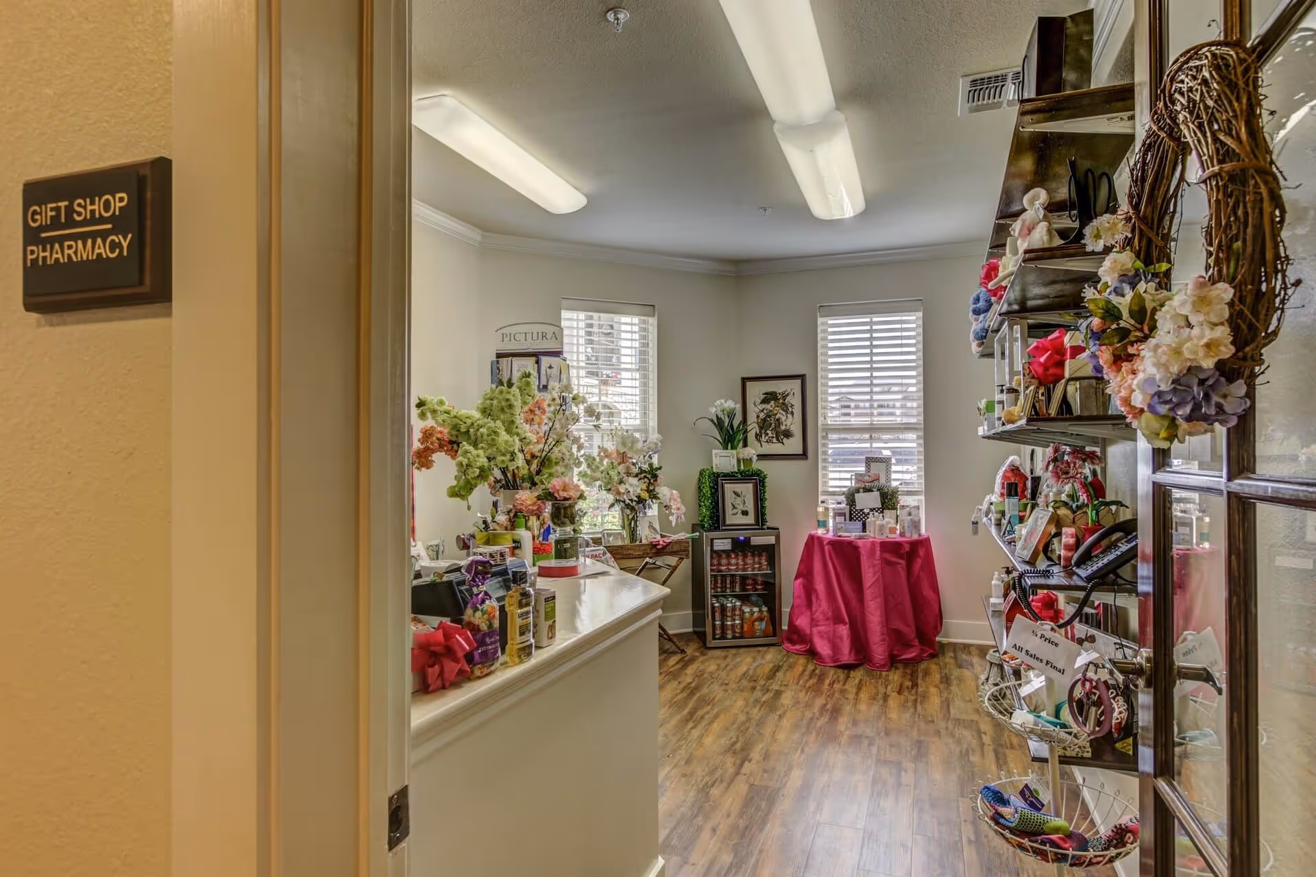 Interior view of a small gift shop area inside a senior living facility. The room has wooden flooring, two windows with blinds, and shelves filled with various gift items and decorations including flowers and wreaths. A sign on the wall outside the room reads 'Gift Shop Pharmacy.'