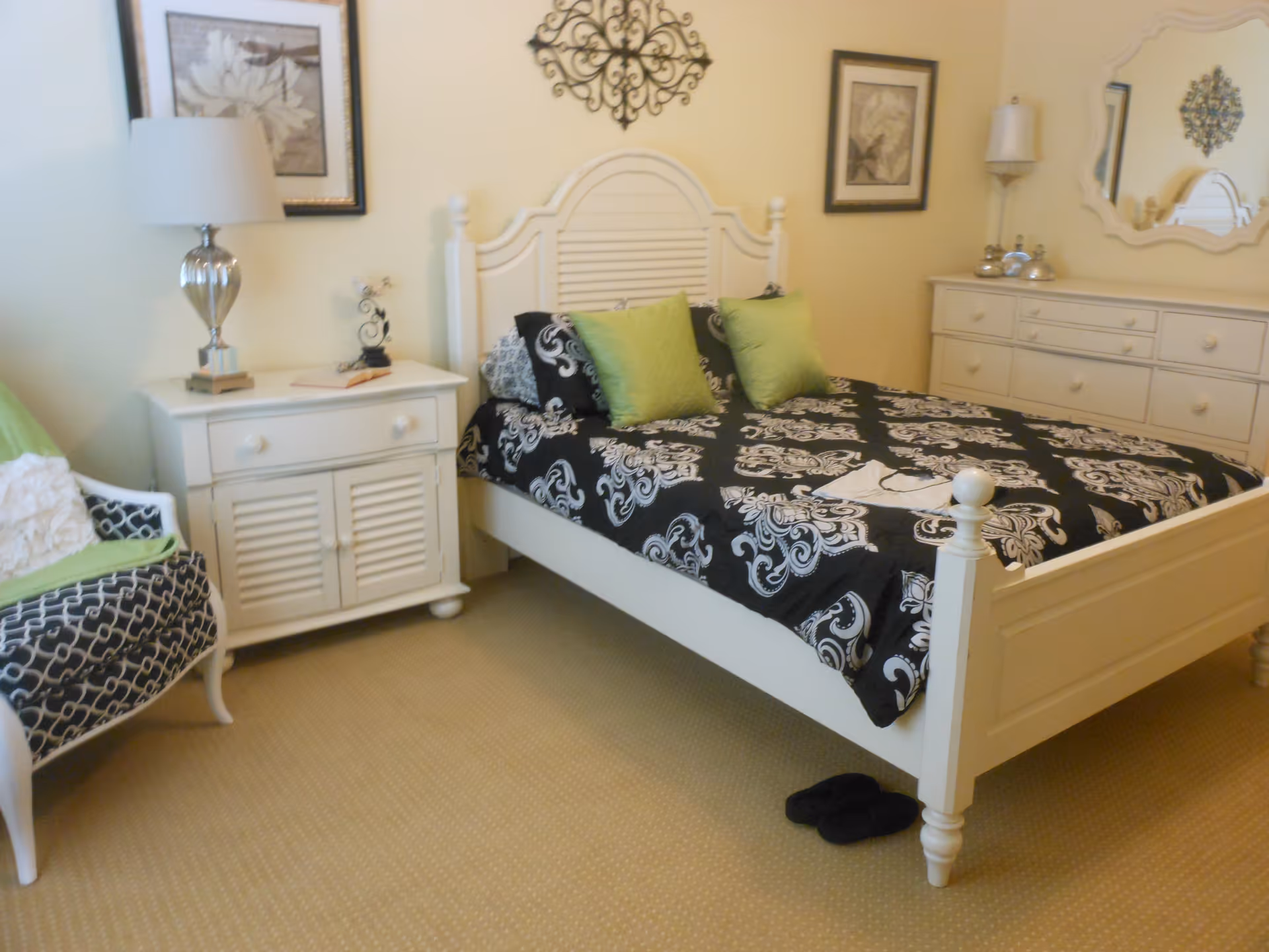 A bedroom with a white wooden bed frame featuring a black and white patterned bedspread and two green pillows. There is a white nightstand with a lamp and decorative items to the left of the bed, and a white dresser with a mirror and lamp on the right side. A patterned armchair with a green throw is positioned near the nightstand. The walls are light-colored and decorated with framed artwork and a decorative metal wall piece above the bed.