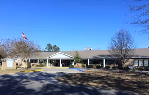 Front view of a single-story senior living center with a flag, trees, and parking.