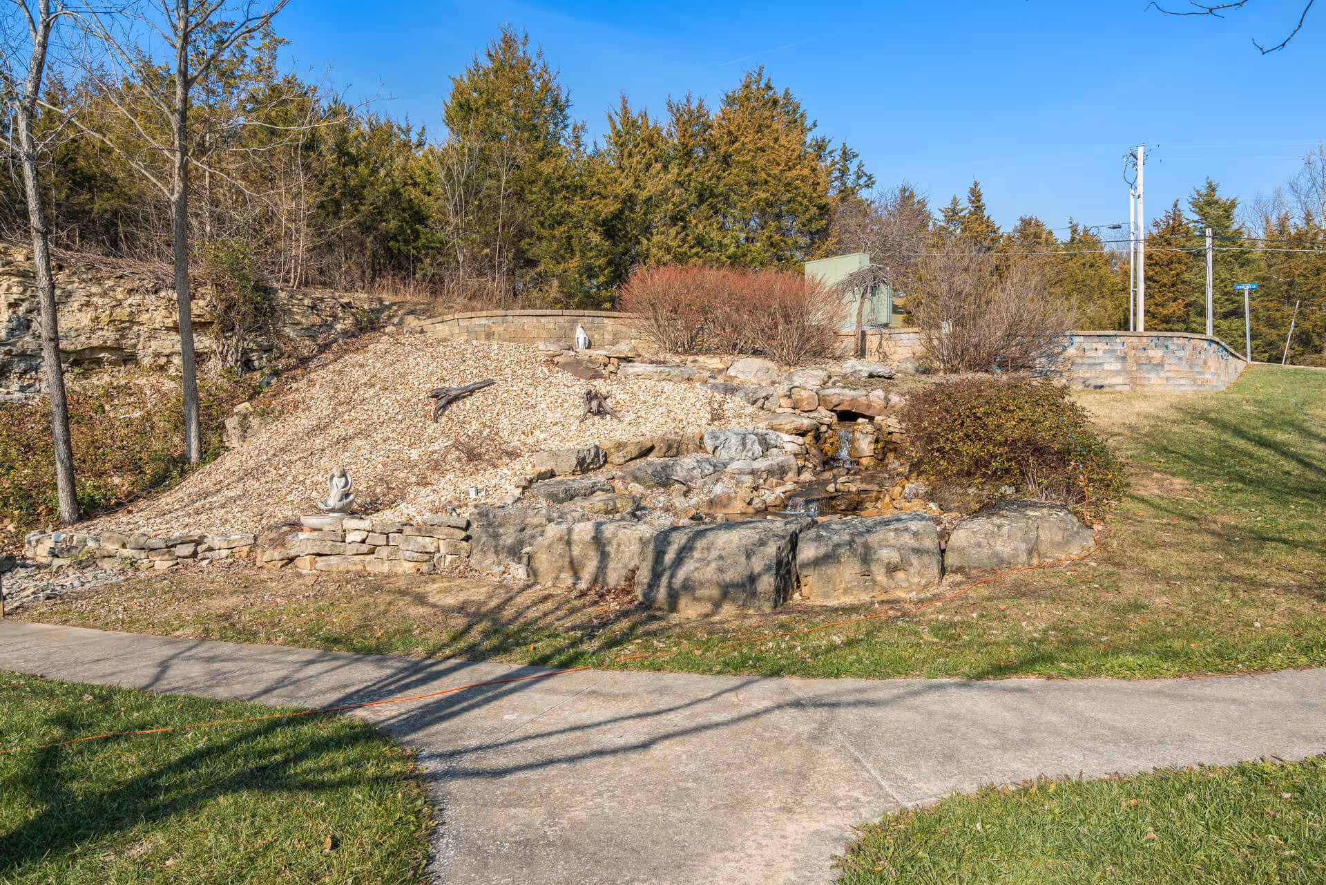 Outdoor landscaped area with a small rock waterfall feature surrounded by bushes and trees, a stone retaining wall, and a concrete pathway in the foreground under a clear blue sky.