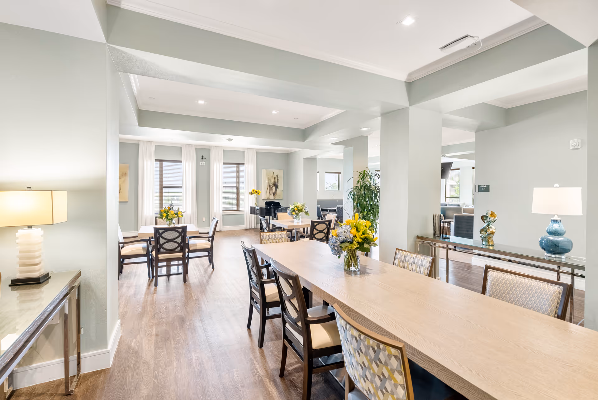 Bright and spacious dining area with multiple tables and chairs arranged neatly. The room features light-colored walls, large windows with white curtains allowing natural light to fill the space, and decorative flower arrangements on the tables. There are modern lamps on side tables and a large plant near the center column, creating a welcoming atmosphere.
