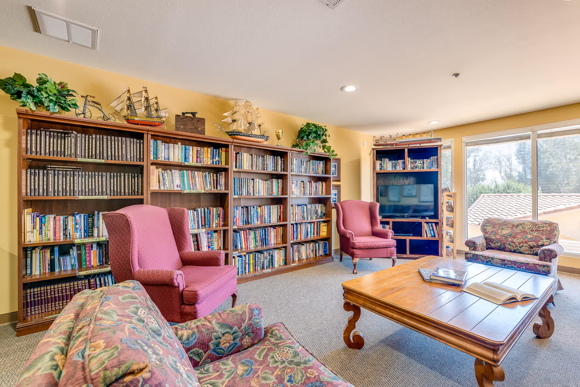 A cozy senior living facility common room with bookshelves filled with books and decorative ship models on top. The room has two pink upholstered armchairs, a floral patterned sofa, a wooden coffee table with books on it, and a TV on a wooden stand near large windows letting in natural light.