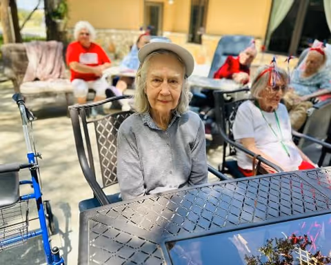 Several elderly residents seated at outdoor patio tables, with a woman in a hat in the foreground.