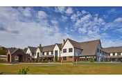 Exterior view of a large senior living facility building under a partly cloudy blue sky, featuring multiple peaked roofs, white walls with brown accents, and a well-maintained lawn in the foreground.